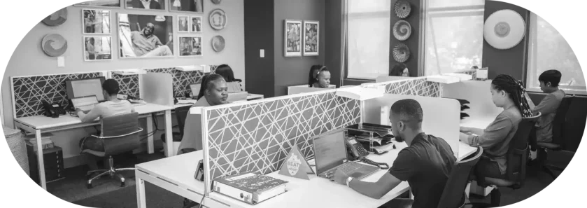 A black and white photo of a people working in an office sitting in front of their laptops.